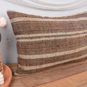 Op de afbeelding: Rechthoekig kussen met bruine en beige strepen. Het kussen is gemaakt van een geweven stof en ligt op een houten oppervlak. Dit is een item voor in huis.