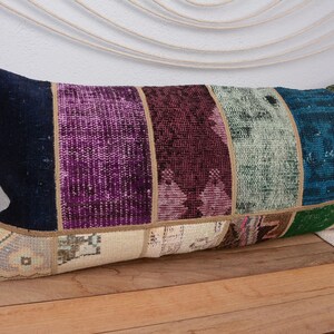Puede incluir: Cojín decorativo rectangular con diseño de patchwork. El cojín presenta un extremo azul marino, seguido de franjas verticales moradas, burdeos, turquesas y verdes. La base del cojín es de color beige con un diseño estampado.