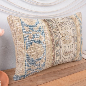 May include: Rectangular decorative pillow with a vintage-style design. The pillow features a beige background with blue and brown patterned stripes. The pillow is placed on a wooden surface.