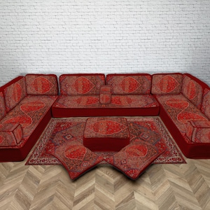 Puede incluir: Un sofá seccional rojo con cojines estampados y una alfombra a juego. El sofá tiene un perfil bajo y está dispuesto en forma de U. La alfombra y los cojines presentan un diseño detallado y ornamentado en tonos rojos, marrones y dorados.
