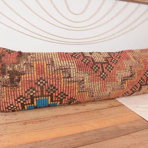 May include: A long, rectangular pillow with a woven, patterned design. The pillow features a mix of colors, including red, brown, yellow, and blue. The pillow is placed on a wooden surface.