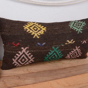 May include: A rectangular, dark brown pillow with colorful geometric patterns in yellow, green, and pink. The pillow is placed on a wooden surface, with a vase of flowers and a decorative basket in the background.