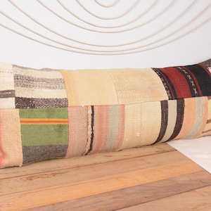 May include: A long, rectangular cushion with a patchwork design. The cushion features various fabric patches in beige, brown, red, and green. The cushion is placed on a wooden surface, with a white background and decorative elements.