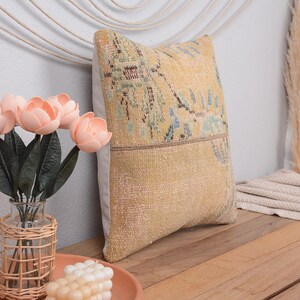 May include: A decorative pillow with a yellow and beige patterned design. The pillow is rectangular and has a textured appearance. A vase of pink flowers and a candle are on a wooden surface.