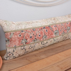 May include: Rectangular decorative pillow with a vintage rug design. The pillow features a central band of red and blue patterns, framed by beige and cream-colored sections. The pillow is placed on a wooden surface.