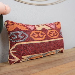 Peut inclure: Un coussin rectangulaire avec un motif géométrique coloré dans des tons de rouge, orange, jaune et blanc. Le coussin est fait d'un tissu tissé et a une bordure frangée.