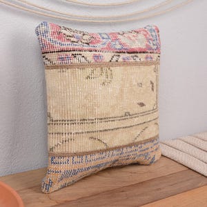 May include: Rectangular decorative pillow with a beige, blue, and pink patterned design. The pillow has a woven texture and is placed on a wooden surface. The pillow's design includes geometric and floral elements.
