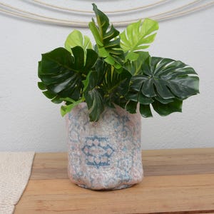 May include: A decorative vase with a light blue and pink pattern holds a vibrant arrangement of green tropical leaves. The vase sits on a wooden surface, creating a natural and inviting aesthetic. The leaves have a glossy finish.