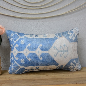 Puede incluir: Una almohada de tiro azul y blanco con un diseño geométrico. La almohada está sobre una superficie de madera con un ramo de flores rosas en un jarrón de mimbre.