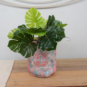 May include: A decorative vase with a floral pattern in red, blue, and white holds vibrant green artificial leaves. The vase sits on a wooden surface, with a neutral-colored runner on the left.