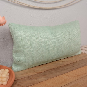 May include: A light green rectangular pillow with a textured woven fabric. The pillow is sitting on a wooden surface.
