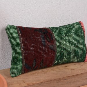 Peut inclure: Coussin rectangulaire décoratif avec un motif patchwork. Le coussin présente une section centrale rouge foncé avec des accents sombres, flanquée de sections vertes à motifs. Les bords sont finis avec une bordure verte et une touche de rose.