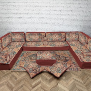May include: A U-shaped sectional sofa with a matching rug and pillows. The sofa and pillows have a patterned design in shades of blue, orange, and beige. The sofa's base is a deep red color. The rug is rectangular.