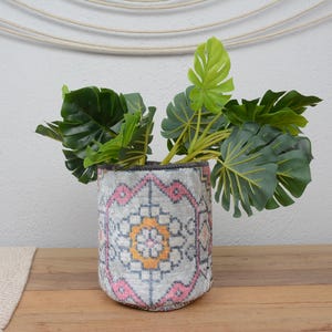 Può includere: Un vaso decorativo cilindrico con un motivo grigio, rosa e arancione. Il vaso è riempito con grandi foglie tropicali verdi. Il vaso è appoggiato su una superficie di legno.