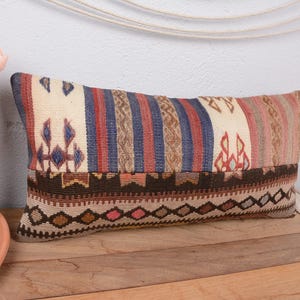 May include: Rectangular decorative pillow with a woven design. The pillow features a cream-colored background with vertical stripes in red, blue, and brown. The bottom section has a geometric pattern in brown, beige, and pink.