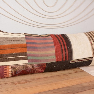 Könnte beinhalten: Ein langes, rechteckiges Kissen mit einem Patchwork-Design. Das Kissen hat verschiedene Abschnitte mit unterschiedlichen Mustern und Farben, darunter Streifen in Orange, Rot und Braun sowie einen cremefarbenen Abschnitt. Das Kissen liegt auf einer Holzoberfläche.