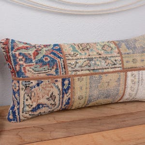 May include: Rectangular patchwork pillow with a vintage rug design. The pillow features a mix of blue, red, beige, and cream colors. The pillow is placed on a wooden surface.