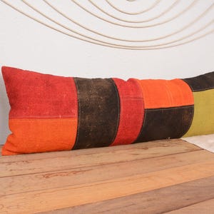 May include: A rectangular decorative pillow with a patchwork design. The pillow features sections of red, orange, brown, and olive green fabric. The pillow is placed on a wooden surface.