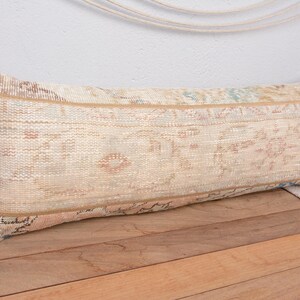 May include: Rectangular beige lumbar pillow with a vintage, patterned design. The pillow features a mix of neutral colors, including beige, tan, and hints of blue and pink. It rests on a wooden surface, suggesting a rustic or bohemian style.