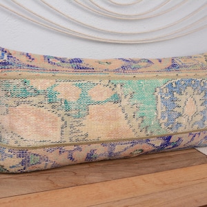 Op de afbeelding: Rechthoekig decoratief kussen met een vintage tapijtontwerp. Het kussen heeft een beige, blauw en teal kleurenpalet met een distressed patroon. Het kussen ligt op een houten oppervlak.