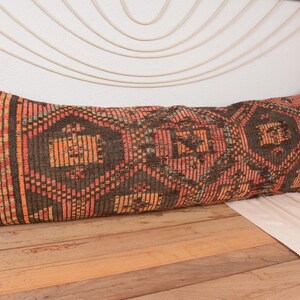 May include: A long, rectangular pillow with a geometric pattern in shades of brown, orange, and red. The pillow is placed on a wooden surface, and the design features diamond and rectangular shapes.