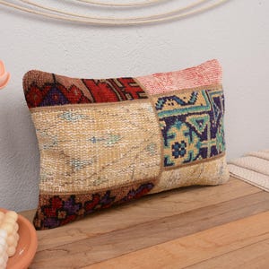 May include: A rectangular throw pillow with a patchwork design. The pillow is made of different fabrics with various patterns in shades of red, blue, green, and beige. The pillow is lying on a wooden surface.