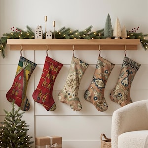 Peut inclure: Cinq chaussettes de Noël suspendues à une étagère en bois, chacune présentant un motif tissé unique dans des couleurs variées, notamment rouge, vert, jaune, beige et bleu. Les chaussettes sont suspendues par des boucles en cuir sur des crochets.