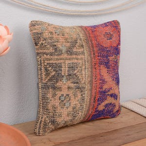 May include: A square decorative pillow with a patchwork design. The pillow features a beige and dark gray patterned section, and a section with a red and purple floral pattern. The pillow is placed on a wooden surface.