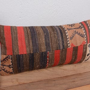 May include: Rectangular pillow with a patchwork design. The pillow features vertical stripes in red, brown, and black, with a tan patterned section on one end. The pillow is placed on a wooden surface.