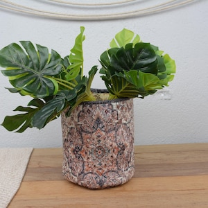 Könnte beinhalten: Eine dekorative zylindrische Vase mit einem detaillierten Muster in Braun-, Orange- und Blautönen. Die Vase enthält mehrere große, leuchtend grüne Blätter. Die Vase steht auf einer Holzoberfläche.