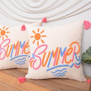 May include: Two white throw pillows with pink tassels. The pillows have the word "Summer" embroidered in pink, orange, and blue with a sun and waves in the background.
