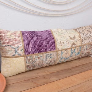 May include: Rectangular patchwork pillow with a mix of colors including purple, beige, and light blue. The pillow is made of various fabric pieces sewn together. The pillow is placed on a wooden surface.