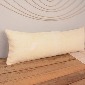 May include: A long, rectangular cream-colored pillow with a textured surface. The pillow is placed on a wooden surface. Decorative elements, including a floral arrangement and a wall hanging, are visible in the background.