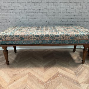 May include: A wooden bench with a patterned fabric cushion. The cushion has a blue, pink, and white floral design. The bench has turned legs and is sitting on a wood floor.