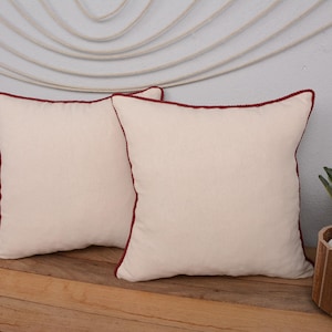 May include: Two square cream-colored throw pillows with a dark red trim. The pillows are placed on a wooden surface. A small potted plant with green leaves sits to the right of the pillows. The background is a light gray wall.