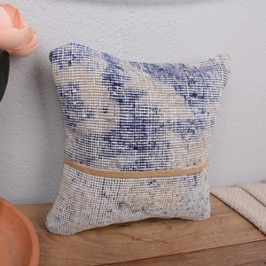 Può includere: Cuscino decorativo quadrato con una trama tessuta nei toni del blu, beige e bianco. Una fascia beige orizzontale accentua la parte inferiore. Il cuscino è posto su una superficie di legno, con un vaso di fiori rosa sullo sfondo.