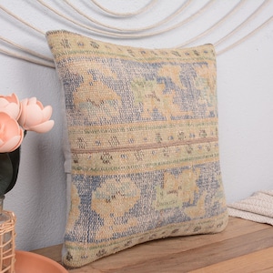 May include: A square decorative pillow with a vintage-style design. The pillow features a beige, blue, and tan pattern. The pillow is placed on a wooden surface, with a vase of pink flowers in the foreground.