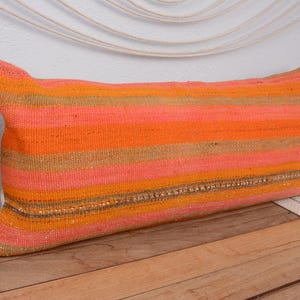 May include: Rectangular decorative pillow with horizontal stripes in shades of orange, peach, and tan. The pillow is made of woven fabric and has a textured appearance. A decorative accent of small beads runs along the center of the pillow.