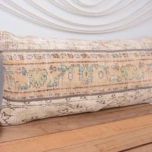 May include: Rectangular beige pillow with a faded, patterned design in shades of blue, green, and brown. The pillow has a distressed, vintage look, with a woven texture. The pillow is placed on a wooden surface.