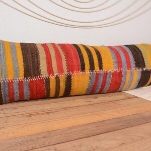 May include: A long, rectangular cushion with a patchwork design. It features vertical stripes in red, yellow, brown, and grey. The cushion is placed on a wooden surface, highlighting its vibrant colours and textured pattern.