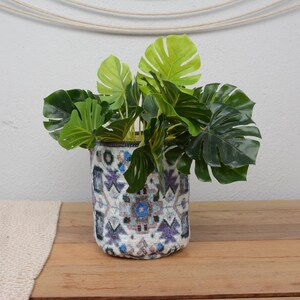 Puede incluir: Una maceta cilíndrica decorativa con base blanca y un patrón geométrico en azul, morado y turquesa contiene vibrantes hojas verdes de Monstera. La maceta está sobre una superficie de madera.