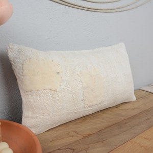 May include: White throw pillow with a textured woven fabric and two off-white fabric patches. The pillow is on a wooden surface.