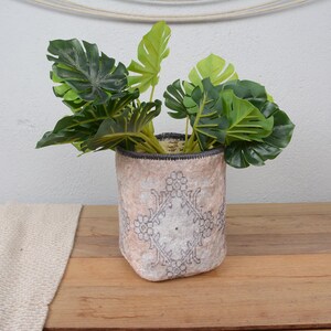 Puede incluir: Una maceta decorativa con un diseño floral. La maceta es de color beige claro con un patrón floral gris oscuro. Contiene varias plantas de hojas verdes. La maceta está sobre una superficie de madera.
