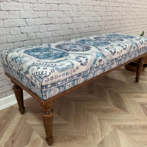May include: A wooden bench with turned legs and a blue and white patterned fabric top. The bench is sitting on a wood floor with a herringbone pattern.