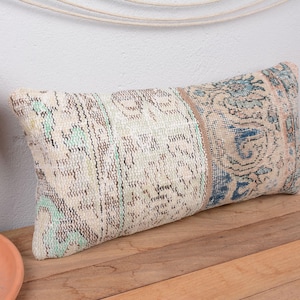 May include: Rectangular decorative pillow with a vintage, distressed look. The pillow features a beige background with intricate patterns in shades of blue, green, and brown. The pillow is placed on a wooden surface.