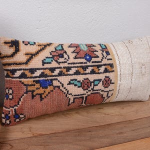 May include: Rectangular decorative pillow with a patterned design. The pillow features a beige background with a central section of brown, blue, and red floral motifs. The right side is a solid beige color. The pillow is placed on a wooden surface.