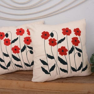 May include: Two white cotton pillow covers with red embroidered poppy flowers. The pillows are on a wooden surface.