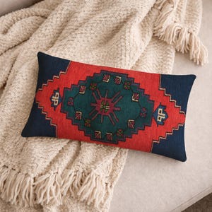 May include: Rectangular decorative pillow with a geometric pattern in red, teal, and navy blue. The pillow is placed on a cream-colored blanket with fringe. The pillow's design features a central teal diamond shape with a red and navy blue border.