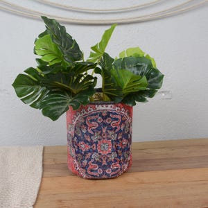Puede incluir: Una maceta decorativa con un patrón detallado y colorido, que contiene una planta verde vibrante con hojas grandes. La maceta presenta una base roja y un diseño azul y crema. La maceta está sobre una superficie de madera.