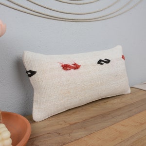 May include: A white rectangular pillow with black and red embroidered designs. The pillow is on a wooden surface.